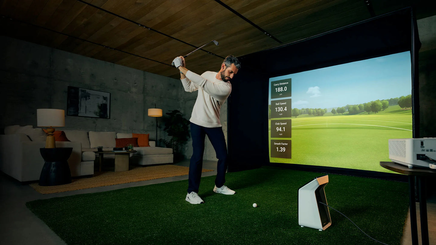 Man using Garmin Approach R50 golf simulator in a home setting with a projection screen displaying swing metrics.