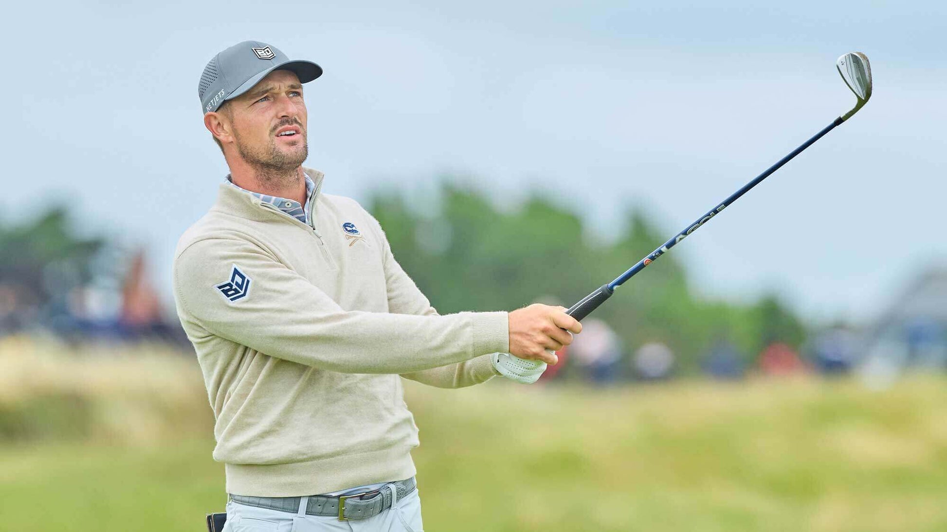 Golfer swinging a club, wearing a gray lacoste sweater and cap in a green outdoor setting.