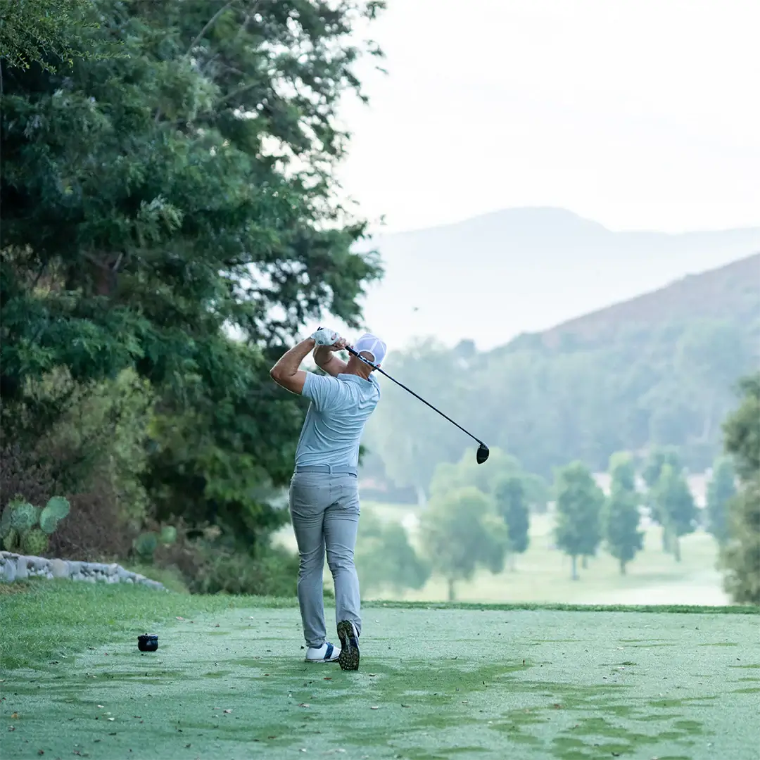 A golfer swings his driver on a picturesque golf course surrounded by trees and hills.